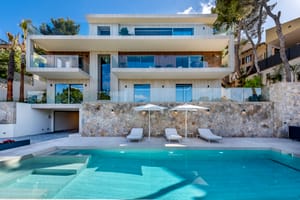 Three-story modern villa with glass railings, stone retaining wall, swimming pool, and sun loungers on a paved terrace.