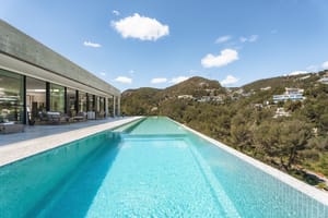 A long turquoise infinity pool runs alongside a modern villa with glass walls, overlooking a verdant mountain range.