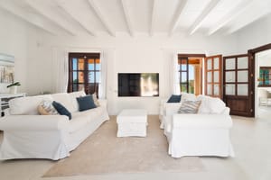 Salón con sofás blancos, otomana central y vigas de techo pintadas de blanco.