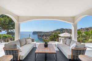 A covered outdoor lounge featuring gray sofas and wooden tables under white arches, overlooking the blue sea and cliffs.