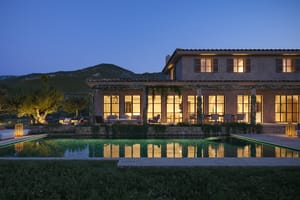 Exterior view of a stone finca at dusk, featuring a rectangular swimming pool, illuminated ground floor windows, and surrounding mountain silhouettes.