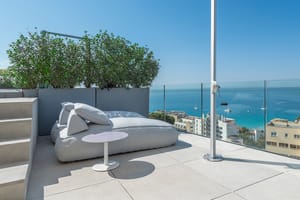 A rooftop terrace featuring a large gray outdoor daybed, a white side table, and a glass balustrade overlooking the sea.