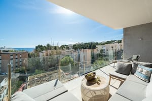 A covered terrace with grey outdoor sofas, a circular wooden coffee table, and glass railings overlooking a coastal town.