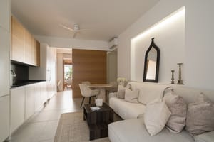 A bright apartment living space featuring a white sofa, minimalist kitchenette with wood cabinets, and dining area.