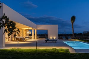 Exterior view of a modern finca at twilight, featuring a lit swimming pool, stone terrace, and warm architectural lighting.