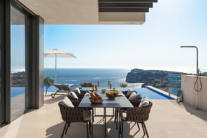 Covered outdoor dining terrace with a dark metal table, woven chairs, and an infinity pool edge against a Mediterranean sea view.