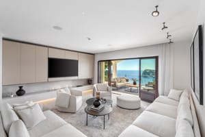 Modern white living room with soft seating and circular tables facing wide glass doors and a coastal view.