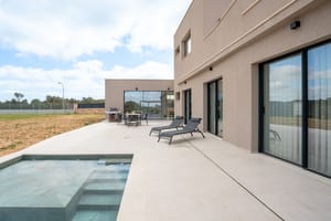 Wide view of a modern villa terrace featuring a rectangular swimming pool, sun loungers, and large sliding glass doors.