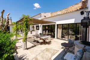 A sunny stone-paved terrace with a wooden pergola covering a dining table and chairs, adjacent to a white house facade.