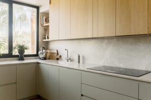 Küche mit salbeifarbenen Unterschränken, oberen Eichenelementen und Steinplatten, durch Tageslicht beleuchtet.