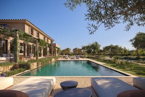 A wide exterior view of a finca with a rectangular swimming pool, gravel patio, sun loungers, and pergola overlooking mountains.