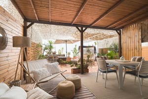 A shaded outdoor terrace with wooden panel walls, dining furniture, a sofa, and potted plants under a perforated roof.
