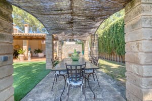 A stone-columned patio with a trellis roof and iron dining set, overlooking a green garden and building exterior.