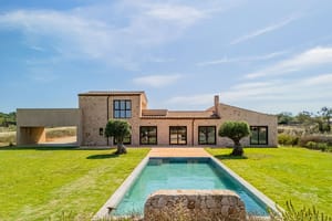 Exterior view of a stone finca in Santanyí featuring a rectangular swimming pool, manicured lawn, and large glass doors.