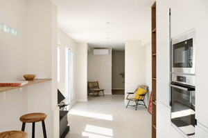 Interior view from a kitchen looking towards a living area with white walls, tiled floors, a stool, and a yellow chair.