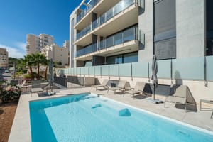 A rectangular outdoor swimming pool with light blue water sits on a stone terrace in front of a modern multi-story apartment building with glass balcony railings.