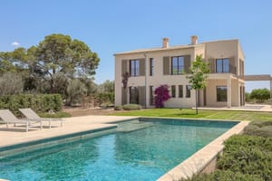 Exterior view of a beige stone finca with wooden shutters, a swimming pool, and lounge chairs in a landscaped garden.