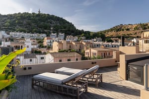 Rooftop terrace featuring two wooden sun loungers with white cushions, overlooking a hillside town in Genova under clear skies.
