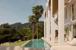 Rectangular turquoise lap pool beside a white modern villa with wooden pergolas, palm trees, and mountain views.