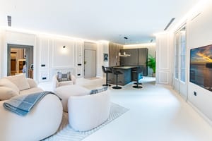 Wide shot of a modern white open-plan living room with a kitchen island, textured armchairs, and floor-to-ceiling windows.