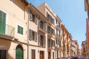 Multi-story Mediterranean townhouses featuring sand-colored plaster, green wooden shutters, and wrought-iron balconies.
