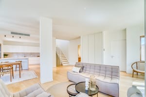 Modern open-plan living room with light oak flooring, grey tufted sofas, and a white architectural staircase.