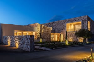 Exterior view of a modern stone-clad finca in Santanyí at twilight, showing illuminated interior spaces and landscaped grounds.