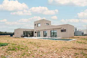 Exterior view of a modern two-story beige villa with a swimming pool, flat roof, and large glass sliding doors.