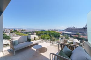Terraza amplia con sofás grises de exterior y cojines verdes, suelo de piedra y vistas al puerto y al mar.