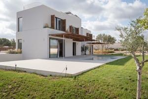 Modern two-story white villa with wooden shutters, a large pergola, and stone terrace overlooking a green lawn.