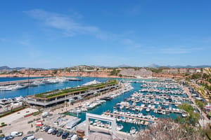 Elevated view overlooking Port Adriano marina with docked yachts, a blue sea bay, and the coastal town in the background.