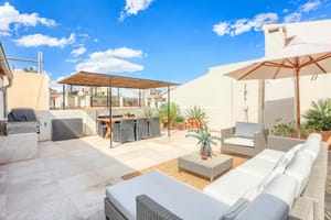 A bright rooftop terrace with a dining area under a wooden pergola and a lounge seating area with white cushions and a large cream umbrella under a clear blue sky.