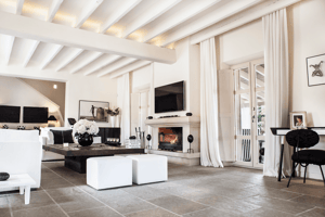 Bright living area with a stone fireplace, exposed beam ceiling, stone flooring, and white linen curtains.