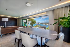 Dining room with a large oval grey stone table and upholstered chairs, facing an open terrace overlooking a blue bay.