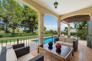 A covered terrace with comfortable outdoor seating overlooks a blue swimming pool and surrounding green gardens on a sunny day.