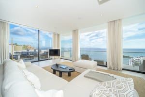 Modern living room featuring floor-to-ceiling glass walls, white sofas, woven area rug, and panoramic coastal views.