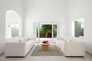 Salón blanco de doble altura con ventana arqueada, dos sofás color crema y puertas acristaladas hacia el jardín