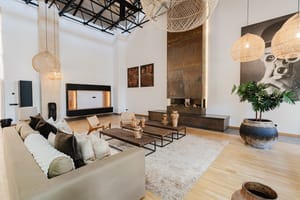 A spacious living area featuring a double-height ceiling with exposed black metal trusses, a tall rusted-metal fireplace surround, and woven pendant lighting.