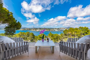 Modern outdoor terrace featuring woven grey furniture, a white coffee table with orchids, and a glass balustrade overlooking a bay with mountains in the background.