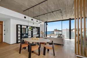 Modern open-plan living and dining room with concrete ceiling, wooden floors, and floor-to-ceiling windows overlooking the sea.