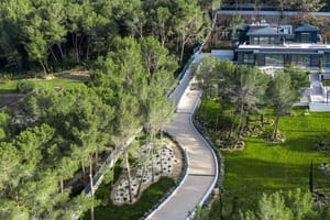 Aerial view of a modern villa with a flat roof, surrounded by gardens, pine trees, and a winding driveway.