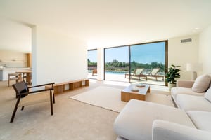Bright open-plan living room featuring neutral sofas, a woven wood armchair, and floor-to-ceiling glass doors opening to a pool deck.
