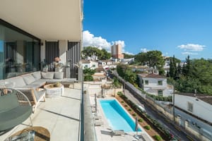 A covered terrace with grey sectional seating overlooks a rectangular swimming pool surrounded by a paved garden in a suburban setting.