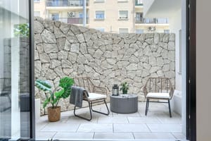 A private outdoor patio featuring a high dry-stone boundary wall, light-toned stone paving, and two rattan chairs with a small table.