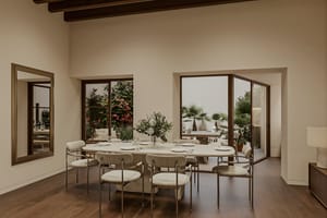 Dining table with eight chairs beside large windows and glazed doors overlooking a planted garden with bougainvillea and palms.