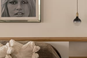 A close-up of a bedroom interior showing a textured brown bedspread, off-white decorative pillows with tassels, and a floating light-oak headboard shelf with a pendant light.
