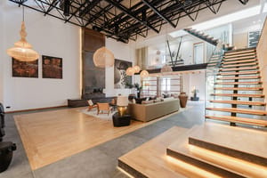 A spacious apartment interior with high ceilings, exposed metal roof trusses, a floating staircase, and neutral-toned furniture.