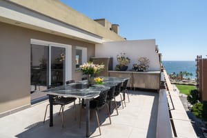 A spacious outdoor terrace with a long dining table, modern chairs, and an open view overlooking the city and the sea.