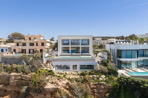 Wide exterior view of a modern white multi-level villa built into a rocky coastline with a swimming pool and glass walls.