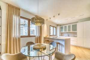 Modern open-plan kitchen with white cabinetry and dining area featuring a round glass table, beige armchairs, and floor-length curtains.